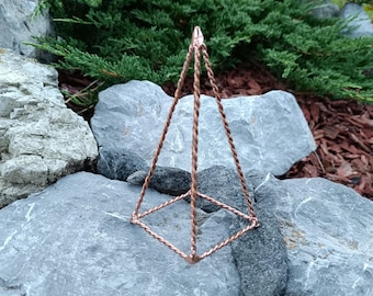 May include: A copper-colored geometric pyramid-shaped hanging ornament. The pyramid is constructed with twisted wire and has a square base. The ornament is displayed outdoors against a backdrop of gray rocks and green foliage.