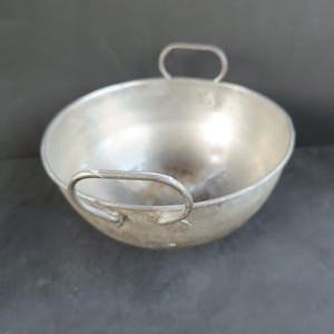 May include: A silver-colored, round cooking pot with two arched handles. The pot has a wide, open bowl shape and appears to be made of metal. The handles are attached to the sides of the pot, and the pot is sitting on a dark surface.