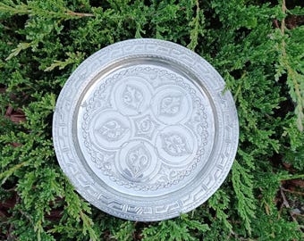 Peut inclure: Une assiette ronde en métal argenté avec un motif floral en relief complexe au centre. L'assiette a une bordure décorative et est présentée sur un lit de feuillage vert. Diamètre d'environ 20 cm.