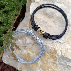 May include: Two braided rope bracelets. One is black with a gold-colored metal accent, and the other is light gray with a silver-colored metal accent. The bracelets are displayed on a light-colored stone surface.