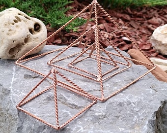 Op de afbeelding: Vier koperkleurige draadpiramides van verschillende afmetingen zijn gerangschikt op een grijs stenen oppervlak. De piramides hebben een gedraaid draadontwerp, waardoor een geometrisch en decoratief effect ontstaat. De achtergrond omvat groen en stenen.