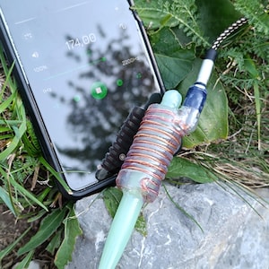 Peut inclure: Gros plan d'une antenne enroulée de couleur verte et cuivre connectée à un smartphone via une prise audio de 3,5 mm. L'écran du téléphone affiche une fréquence de 174,00 Hz et un volume de 100%.