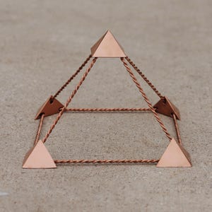 Puede incluir: Una escultura en forma de pirámide de color cobre. La escultura presenta cuatro piezas triangulares de cobre conectadas por alambre de cobre trenzado, formando una forma geométrica tridimensional. La pirámide se encuentra sobre una superficie de tono neutro.