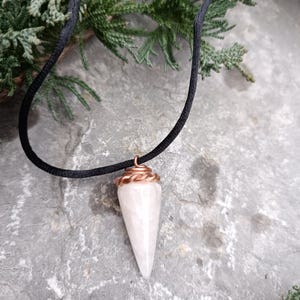 Puede incluir: Un colgante de cristal blanco en forma de punta en un collar de cordón negro. El cristal está envuelto en alambre de cobre en la parte superior.