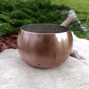 Puede incluir: Una cacerola de color cobre con un cuerpo redondeado y un mango de madera. El interior de la cacerola es de un tono más oscuro de cobre. La cacerola está sobre una superficie de color claro, con follaje verde en el fondo. El mango mide unos 13 cm.