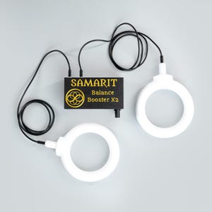 May include: A black device labeled "SAMARIT Balance Booster X2" with gold lettering and a circular logo, connected by black cords to two white, circular devices. The image is set against a light gray background.