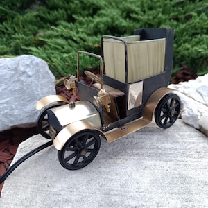 May include: A vintage-style model car with a black body, gold-colored accents, and black wheels. The car has a detailed interior and a fabric top. The model is placed on a light gray stone.