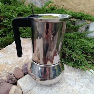 May include: A stainless steel stovetop espresso maker with a black handle. The coffee pot is cylindrical with a rounded base and a lid. The polished metal reflects the surrounding environment. The espresso maker is sitting on a light-colored stone surface.