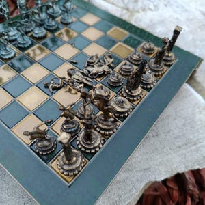 Può includere: Un set di scacchi dettagliato con pezzi color bronzo su una scacchiera a scacchi verde e oro. I pezzi degli scacchi sono finemente progettati, raffiguranti varie figure. La scacchiera è incorniciata da un bordo verde scuro, creando un aspetto classico ed elegante.