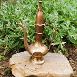 May include: A brass oil lamp with a long curved spout, a slender neck, and a rounded base. The metal surface features intricate etched floral patterns and a hinged lid with a pointed finial. A thin handle is attached to the side.