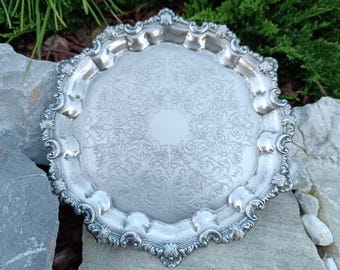 May include: Silver-colored, ornate serving tray with a detailed floral pattern in the center. The tray has a scalloped edge with decorative accents. The tray is round and appears to be made of metal.