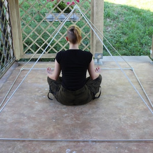 May include: A person sits in a meditative pose inside a white, string-based pyramid structure. The pyramid is set up on a concrete patio.
