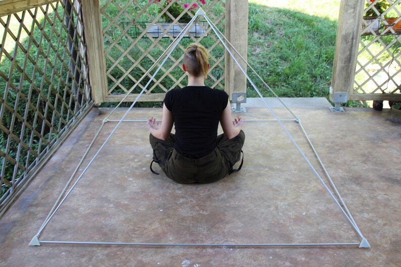 May include: A person sits in a meditative pose inside a white, string-based pyramid structure. The pyramid is set up on a concrete patio.