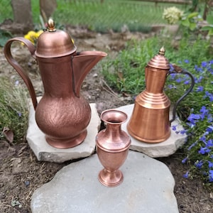 Könnte beinhalten: Ein Set aus drei Kupfergefäßen, bestehend aus einer großen Kanne mit Deckel und geschwungenem Griff, einer kleineren Kanne mit Deckel und dunklem Metallgriff sowie einer kleinen offenen Vase, alle mit metallischem Glanz auf Steinen.