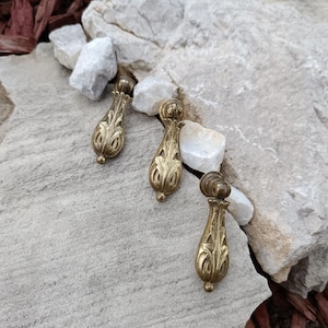 Może przedstawiać: Trzy antyczne mosiężne uchwyty do szuflad z ozdobnymi, kwiatowymi wzorami. Każdy uchwyt ma kształt łzy z dekoracyjną górą i dołem. Uchwyty są ułożone na szarej kamiennej powierzchni.