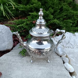 Puede incluir: Pequeña tetera plateada ornamentada, con tapa y asa decorativas. La tetera presenta intrincados diseños florales y se apoya sobre tres pequeñas patas. El pico es curvo y el asa tiene un diseño abierto único. La tetera mide unos 15 cm de altura.