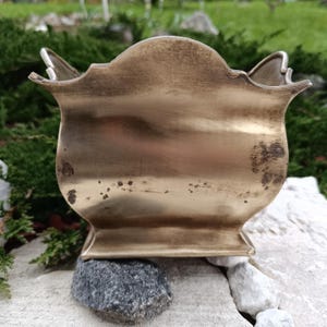 May include: A vintage brass planter with a decorative, scalloped rim. The planter has a slightly aged appearance with some dark spots, suggesting it is an antique. The base is square and it sits on a small rock.