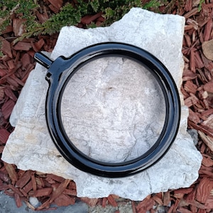 Puede incluir: Un anillo de cortina negro brillante con un pequeño accesorio saliente. El anillo es circular y parece ser de metal. Descansa sobre una piedra gris clara, con mantillo marrón y follaje verde en el fondo.