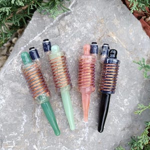 May include: Five glass pipes with different colored glass and copper wire coils. The pipes are on a gray rock surface.