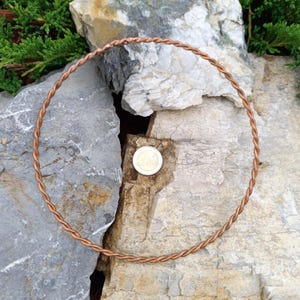 Puede incluir: Un círculo de alambre trenzado de color cobre, posiblemente un collar o un elemento decorativo, descansa sobre una superficie de piedra natural. Se coloca una moneda dentro del círculo como referencia de tamaño. El alambre tiene una apariencia trenzada.
