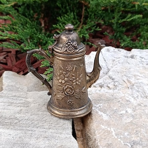 Könnte beinhalten: Kleine Teekanne aus Metall im antiken Stil mit aufwendigen Blumen- und Beerendesigns. Die Teekanne hat einen Klappdeckel, einen gebogenen Ausguss und einen Griff. Die Farbe ist ein angelaufenes Bronze, auf einer grauen Steinoberfläche.