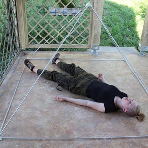 May include: A person lying on their back inside a white pyramid structure made of string. The pyramid is on a concrete floor.