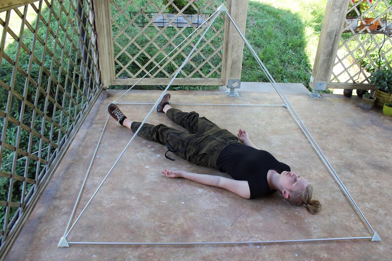 May include: A person lying on their back inside a white pyramid structure made of string. The pyramid is on a concrete floor.