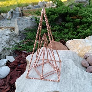 May include: A copper-colored geometric hanging planter with a pyramid design. The planter is made of twisted wire and is suspended by three chains. The planter is sitting on a large, light-colored rock, with greenery in the background.