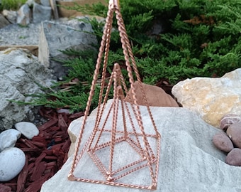 Op de afbeelding: Een koperkleurige geometrische hangende plantenbak met een piramide-ontwerp. De plantenbak is gemaakt van gedraaide draad en wordt opgehangen door drie kettingen. De plantenbak staat op een grote, lichtgekleurde steen, met groen op de achtergrond.