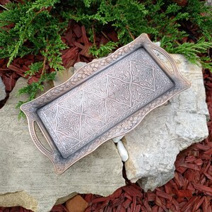 Puede incluir: Una bandeja rectangular de metal con intrincados diseños florales y geométricos. La bandeja tiene un borde y asas de color bronce, con un interior plateado. La bandeja descansa sobre una piedra gris, con vegetación y mantillo rojo en el fondo.