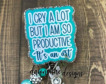 May include: A teal badge reel with the text "I cry a lot but I am so productive. It's an art." The badge has a silver glitter border and a beaded accent with teal and silver beads. The reel has a clear plastic clip.
