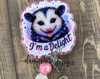 May include: A pink glitter badge holder with a cartoon possum with the text "I'm a Delight" and a clear retractable badge reel with pink, white, and silver beads.