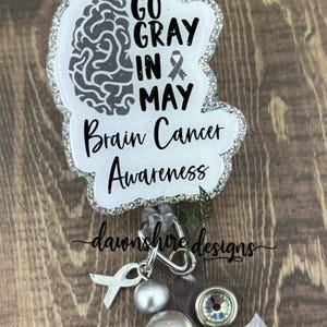 May include: A white and silver glitter brain-shaped badge holder with the text "Go Gray in May Brain Cancer Awareness" and a silver ribbon charm.