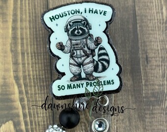 May include: A green and black badge reel with a raccoon wearing an astronaut suit. The badge reel has the text "Houston, I have so many problems".