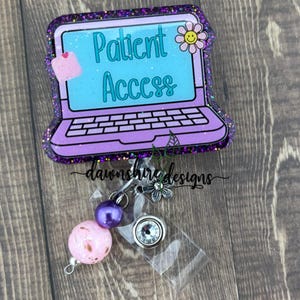 May include: A purple and teal badge reel with the words "Patient Access" on a laptop design. The badge reel has a glittery border, a pink and purple bead, and a silver charm. The design includes a small flower and a note.