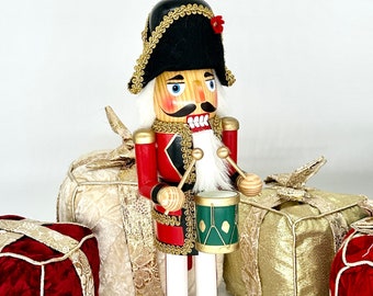 May include: A wooden nutcracker dressed as a soldier, wearing a black hat with gold trim and a red jacket with gold trim, holding drumsticks and a green drum with gold trim. The nutcracker is standing in front of three wrapped gifts, one red, one gold, and one white.