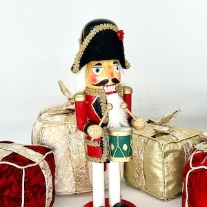 May include: A wooden nutcracker dressed as a soldier, wearing a black hat with gold trim and a red jacket with gold trim, holding drumsticks and a green drum with gold trim. The nutcracker is standing in front of three wrapped gifts, one red, one gold, and one white.
