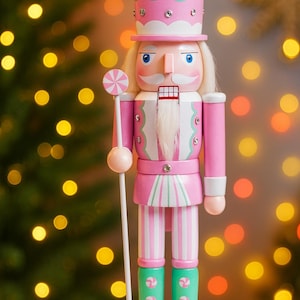 May include: A pink and white wooden nutcracker soldier with a tall hat and a lollipop. The nutcracker has a white mustache and hair, blue eyes, and green boots. The background is blurred with Christmas tree lights.