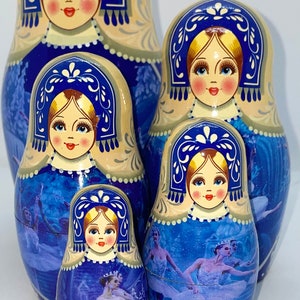 May include: A set of five Russian nesting dolls, or Matryoshka dolls, in varying sizes. Each doll features a painted face and a blue and white design with a ballet theme. The dolls are arranged together.