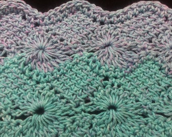 May include: Close-up of a crocheted fabric with a repeating pattern of flowers. The fabric is a mix of light blue and purple yarn.