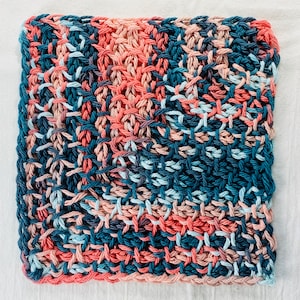 May include: A square knitted dishcloth in shades of blue, pink, and coral. The cloth is made with a textured stitch pattern.