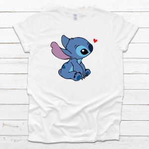 Op de afbeelding: Wit T-shirt met een blauw cartoonfiguur met roze oren en een rood hart erboven. Het figuur zit met gekruiste benen en kijkt naar het hart.