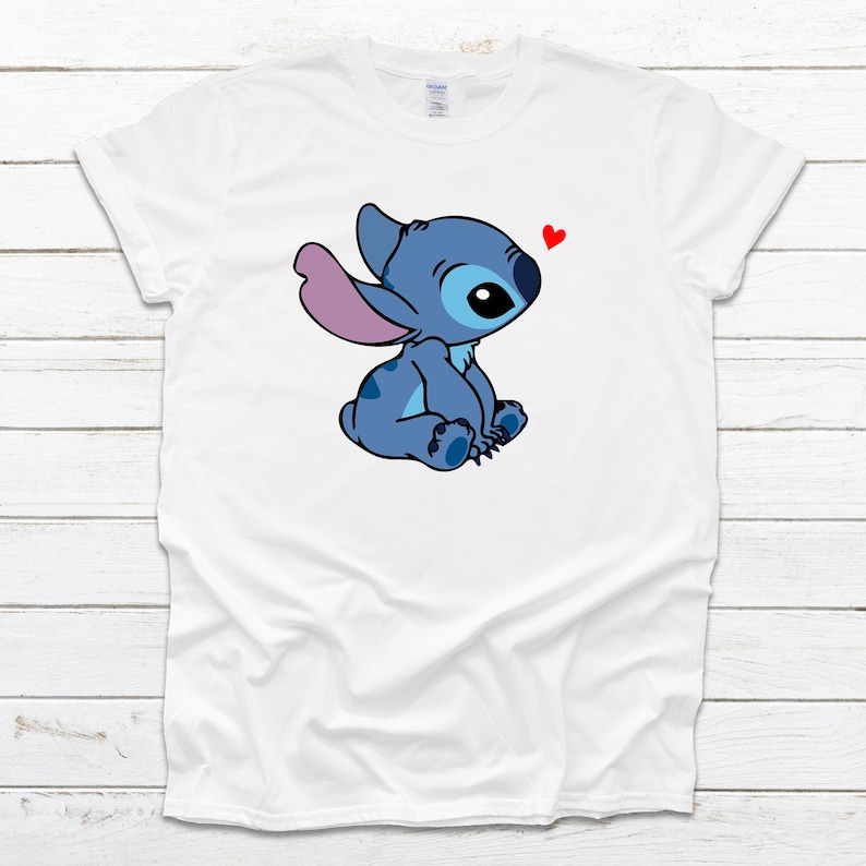 Op de afbeelding: Wit T-shirt met een blauw cartoonfiguur met roze oren en een rood hart erboven. Het figuur zit met gekruiste benen en kijkt naar het hart.