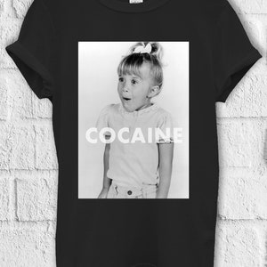 以下が含まれることがあります： 写真の若い女の子が驚いた表情をしている黒のTシャツ。写真の真下に「COCAINE」という文字が白で印刷されています。