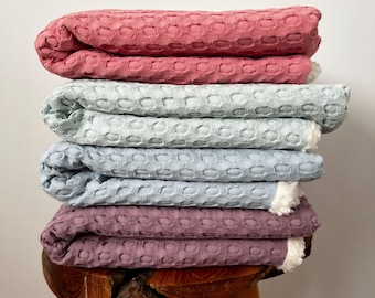 May include: A stack of four waffle weave blankets in pink, light blue, and a darker shade of purple. The blankets are folded and stacked on a wooden stool.