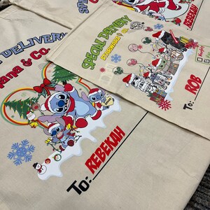 May include: Three white canvas gift bags with colorful designs and the text "Special Delivery" and "Ohana & Co.". The bags have a drawstring closure and are decorated with Christmas-themed characters, including Stitch from Lilo & Stitch, Star Wars characters, and a snowman. The bags have a space for a name and a "To:" label.