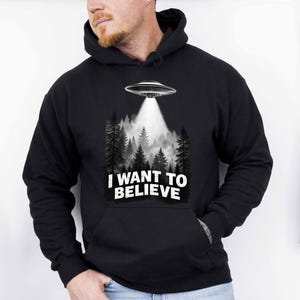 May include: A black hooded sweatshirt featuring a graphic print of a flying saucer hovering over a dark forest with a bright beam of light shining down. The text "I WANT TO BELIEVE" is printed in bold white capital letters below the forest scene.