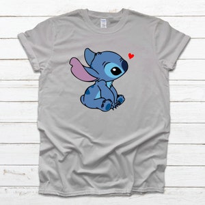 Op de afbeelding: Een grijs T-shirt met een blauw cartoonfiguur met roze oren en een rood hart erboven. Het figuur zit met gekruiste benen en kijkt naar het hart.