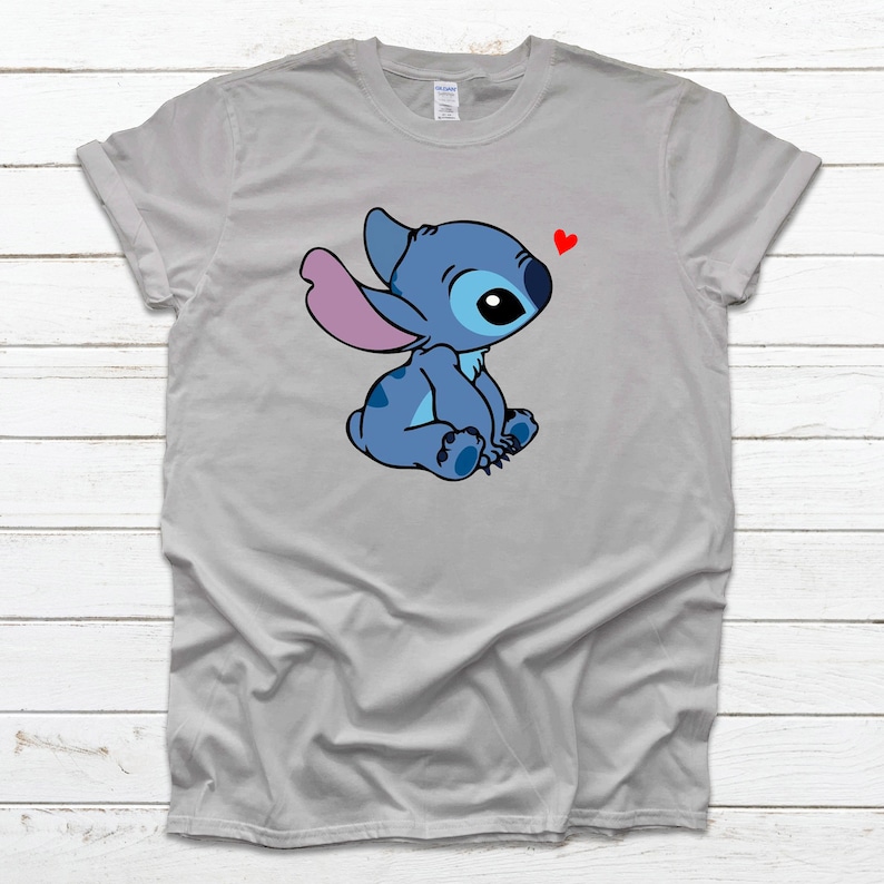 Op de afbeelding: Een grijs T-shirt met een blauw cartoonfiguur met roze oren en een rood hart erboven. Het figuur zit met gekruiste benen en kijkt naar het hart.