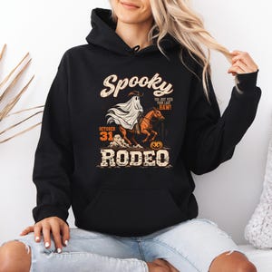 Könnte beinhalten: Ein schwarzer Kapuzenpullover mit einem Grafikdruck eines Geistes mit Cowboyhut auf einem braunen Pferd. Der Text lautet Spooky, You just yee'd your last haw!, October 31 und Rodeo. Das Design enthält einen Kürbis und aufgewirbelten Staub.